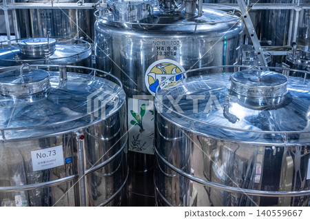 Large shiny stainless steel fermentation storage tanks stand inside spacious modern industrial brewery facility processing liquid commercial beverage products under bright factory lights 140559667