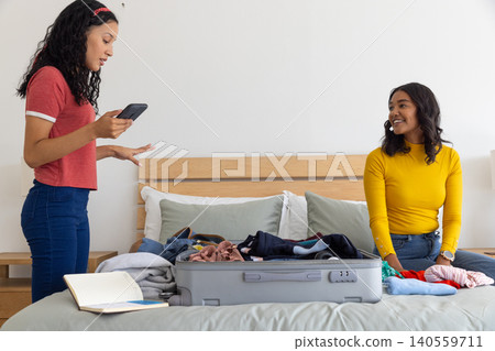 Diverse mom and daughter packing gray suitcase on bed, sorting garments and holding smartphone Diverse mom and daughter packing gray suitcase on bed, sorting garments and holding smartphone 140559711