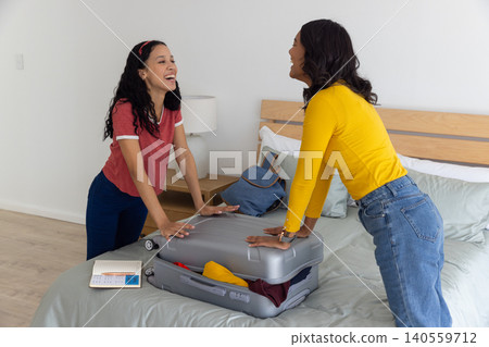 Diverse mom and teenage daughter laughing and leaning forward on bed with open suitcase and planner Diverse mom and teenage daughter laughing and leaning forward on bed with open suitcase and planner 140559712
