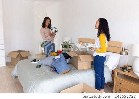 Diverse mom and teenage daughter unpacking moving boxes around bed in bedroom holding soccer ball Diverse mom and teenage daughter unpacking moving boxes around bed in bedroom holding soccer ball 140559722
