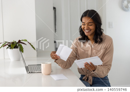 Smiling woman with envelopes and letters at kitchen countertop with laptop mug plant Smiling woman with envelopes and letters at kitchen countertop with laptop mug plant 140559746
