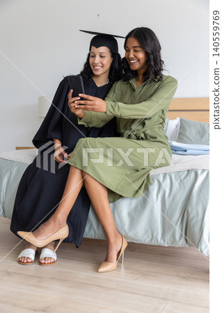 Diverse mom and teenage daughter sitting on bed in bedroom checking smartphone in graduation gown 140559769