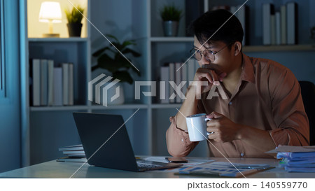 East Asian man focused on work with laptop and mug late at night in office East Asian man focused on work with laptop and mug late at night in office 140559770