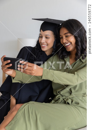 Diverse mom and teenage daughter taking selfie on sofa in graduation gown and jumpsuit Diverse mom and teenage daughter taking selfie on sofa in graduation gown and jumpsuit 140559772