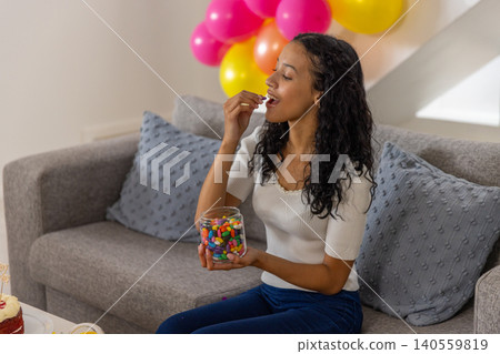 Teenage girl grabbing jellybeans from jar on grey sofa at home with cake and balloons Teenage girl grabbing jellybeans from jar on grey sofa at home with cake and balloons 140559819