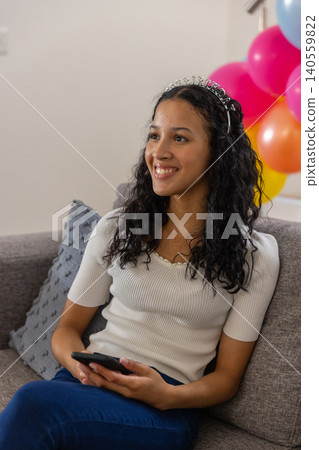 Teenage girl wearing birthday tiara holding smartphone, scrolling on couch with balloons Teenage girl wearing birthday tiara holding smartphone, scrolling on couch with balloons 140559822