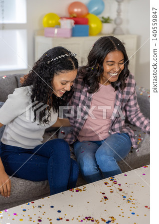 Diverse mom and teenage daughter sitting on sofa in living room with confetti, gift boxes Diverse mom and teenage daughter sitting on sofa in living room with confetti, gift boxes 140559847