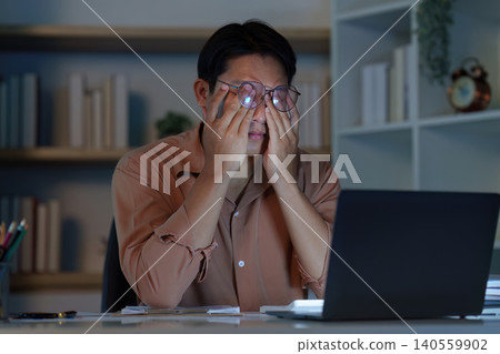 Asian man experiencing eyestrain and fatigue from overwork at remote home office 140559902