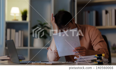 Asian man showing frustration with financial document at remote home office Asian man showing frustration with financial document at remote home office 140560008