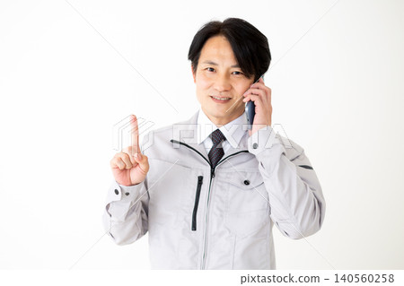 A middle-aged businessman in gray work clothes holds up his index finger while talking on his smartphone against a white background. 140560258