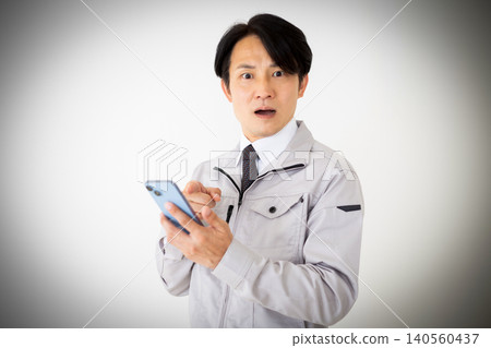 一位身穿灰色工作服的中年商人,在白色背景下看著他的智慧型手機,臉上露出震驚的表情。 一位身穿灰色工作服的中年商人,在白色背景下看著他的智慧型手機,臉上露出震驚的表情。 140560437