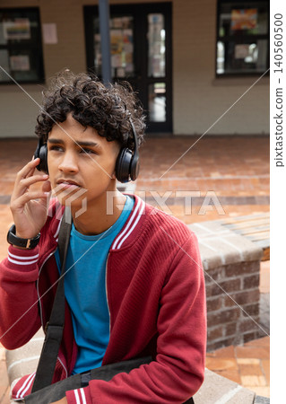 Teenage boy sitting on courtyard planter wearing red jacket adjusting headphones Teenage boy sitting on courtyard planter wearing red jacket adjusting headphones 140560500
