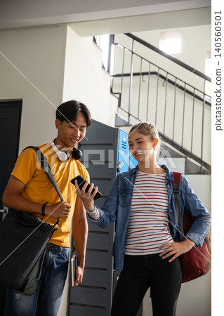 Diverse students comparing smartphone screen, holding notebook in hallway by lockers and stairs Diverse students comparing smartphone screen, holding notebook in hallway by lockers and stairs 140560501