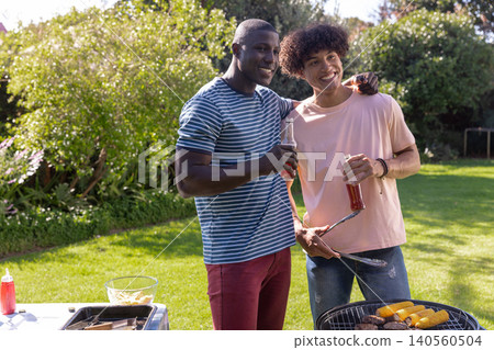 Diverse male friends standing grilling burgers, with glass bottles by charcoal grill in backyard Diverse male friends standing grilling burgers, with glass bottles by charcoal grill in backyard 140560504