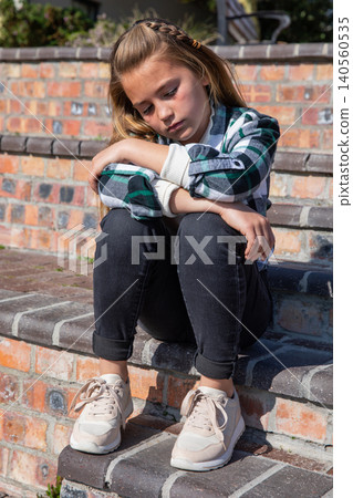 Girl child sitting hugging knees on sunlit front brick steps in plaid shirt, hair clip Girl child sitting hugging knees on sunlit front brick steps in plaid shirt, hair clip 140560535