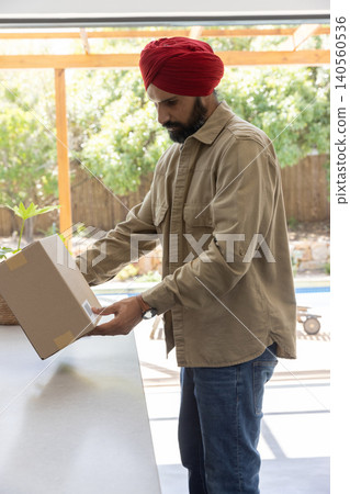 Indian man wearing red turban, shirt examining cardboard box on shaded patio table beside pool Indian man wearing red turban, shirt examining cardboard box on shaded patio table beside pool 140560536