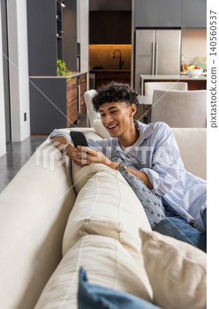 Man reclining on beige sofa in open-plan living kitchen area, checking smartphone 140560537