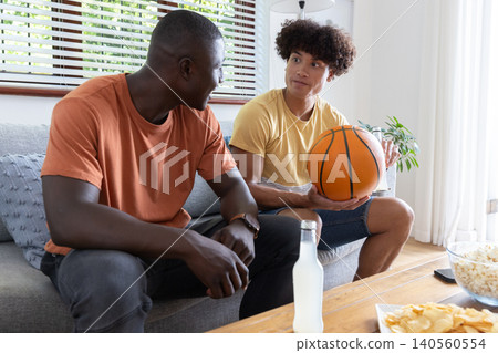 Diverse male friends sitting on sofa in living room holding basketball, grabbing chips Diverse male friends sitting on sofa in living room holding basketball, grabbing chips 140560554