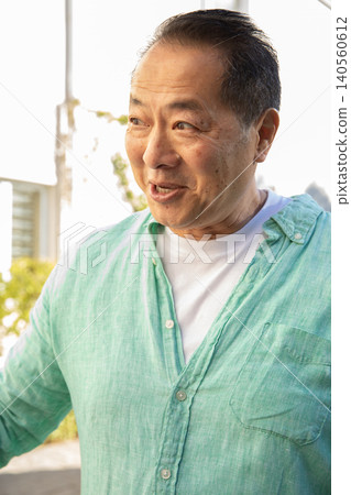 Mature asian man standing on sunny balcony wearing green linen shirt over tee gazing left Mature asian man standing on sunny balcony wearing green linen shirt over tee gazing left 140560612