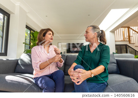 Senior female friends talking on gray sofa in living room with flat-screen television and plant Senior female friends talking on gray sofa in living room with flat-screen television and plant 140560616