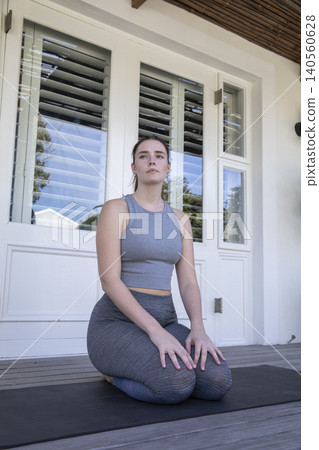 Woman kneeling in grey athletic wear on black yoga mat on wooden porch outside glass doors Woman kneeling in grey athletic wear on black yoga mat on wooden porch outside glass doors 140560628