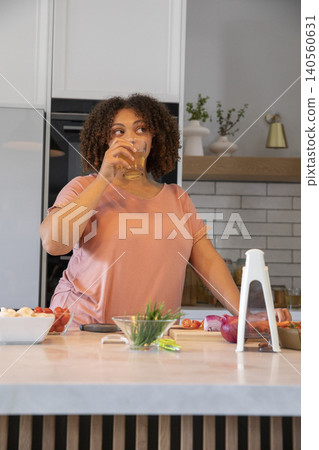 African American woman sipping from glass tumbler while chopping tomatoes on wooden kitchen island African American woman sipping from glass tumbler while chopping tomatoes on wooden kitchen island 140560631