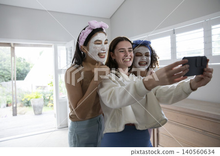Diverse female friends using smartphone for selfie at home wearing facial clay masks and headbands Diverse female friends using smartphone for selfie at home wearing facial clay masks and headbands 140560634