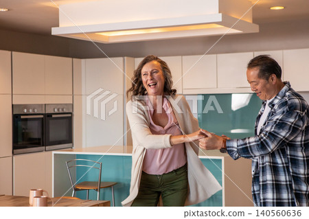 Senior couple dancing hand in hand in modern home with kitchen island and ovens Senior couple dancing hand in hand in modern home with kitchen island and ovens 140560636