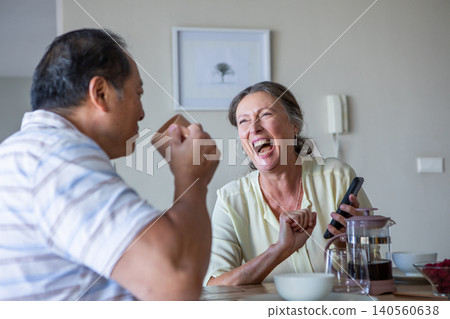 Diverse couple sipping coffee at dining table with french press, laughing with smartphone Diverse couple sipping coffee at dining table with french press, laughing with smartphone 140560638