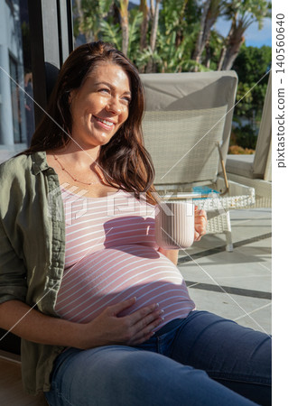 Pregnant woman leaning against glass door holding coffee mug on sunlit patio with wicker furniture 140560640