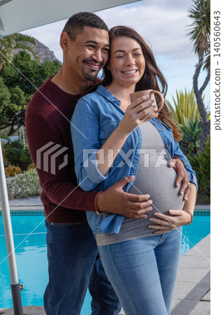 Diverse couple embracing pregnant belly while standing at tiled pool patio holding ceramic mug Diverse couple embracing pregnant belly while standing at tiled pool patio holding ceramic mug 140560643