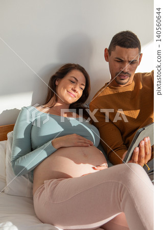 Diverse couple sitting on bed in bedroom against headboard cradling bare pregnant belly with tablet Diverse couple sitting on bed in bedroom against headboard cradling bare pregnant belly with tablet 140560644