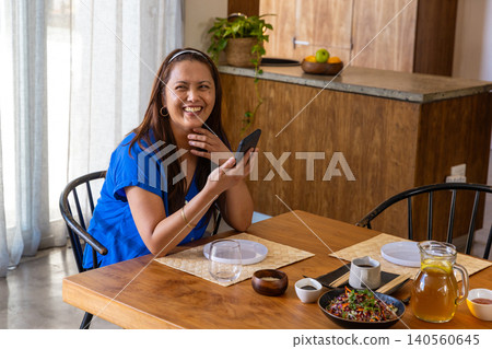 Mature asian woman holding smartphone and resting chin in kitchen at dining table with salad Mature asian woman holding smartphone and resting chin in kitchen at dining table with salad 140560645