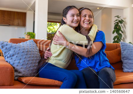 Asian mother and daughter hugging on orange sofa at home with cushions, cabinet with plant Asian mother and daughter hugging on orange sofa at home with cushions, cabinet with plant 140560647