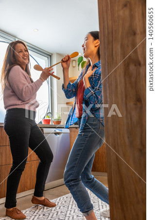 Asian mother and daughter dancing playfully in home kitchen by countertop with spoons Asian mother and daughter dancing playfully in home kitchen by countertop with spoons 140560655
