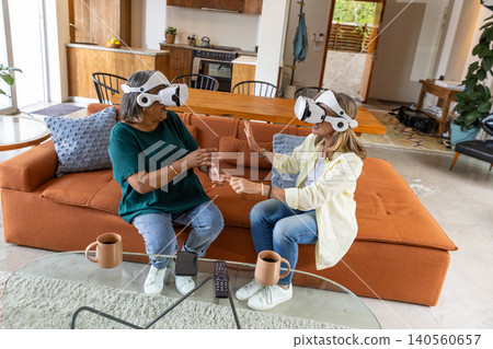Senior female friends sitting on orange sofa wearing vr headsets in living room with mugs Senior female friends sitting on orange sofa wearing vr headsets in living room with mugs 140560657