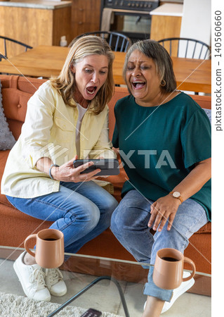 Senior female friends reacting, laughing on couch at home holding smartphone and coffee mugs Senior female friends reacting, laughing on couch at home holding smartphone and coffee mugs 140560660