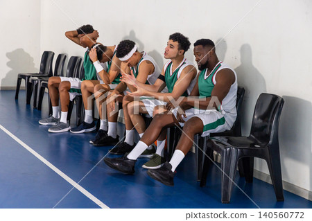 Diverse male basketball teammates sitting on chairs at gym wearing green and white uniforms Diverse male basketball teammates sitting on chairs at gym wearing green and white uniforms 140560772