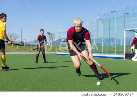 Diverse male field hockey players dribbling ball toward goal cage on turf pitch with sticks Diverse male field hockey players dribbling ball toward goal cage on turf pitch with sticks 140560784