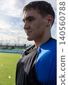 Teenage boy in blue jersey standing on turf field beside lighting poles and track, copy space 140560788