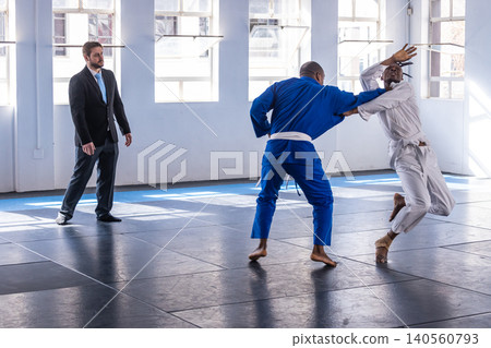 Diverse male sparring fighters in gis exchanging grips on mats in dojo with referee watching Diverse male sparring fighters in gis exchanging grips on mats in dojo with referee watching 140560793