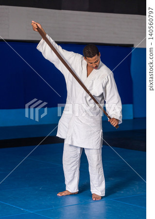 African american man wearing white gi holding brown belt across shoulders in gym with mats African american man wearing white gi holding brown belt across shoulders in gym with mats 140560797