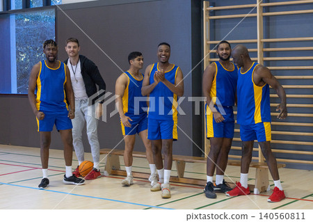Diverse male basketball team wearing matching uniforms on gym court, coach holding whistle Diverse male basketball team wearing matching uniforms on gym court, coach holding whistle 140560811