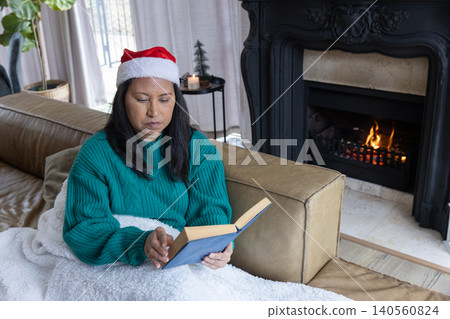 Woman sitting at home on sofa reading hardcover book under plush blanket wearing santa hat Woman sitting at home on sofa reading hardcover book under plush blanket wearing santa hat 140560824
