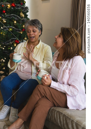Diverse senior women friends chatting over ceramic mugs by decorated Christmas tree in living room 140560856