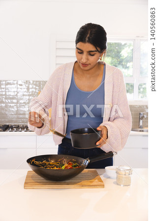 Asian woman ladling noodles and vegetables from black wok into black serving bowl in home kitchen Asian woman ladling noodles and vegetables from black wok into black serving bowl in home kitchen 140560863