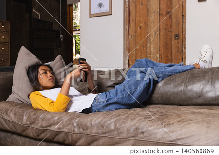 Teenage Asian female reclining on brown sofa with grey pillow in living room, holding smartphone Teenage Asian female reclining on brown sofa with grey pillow in living room, holding smartphone 140560869