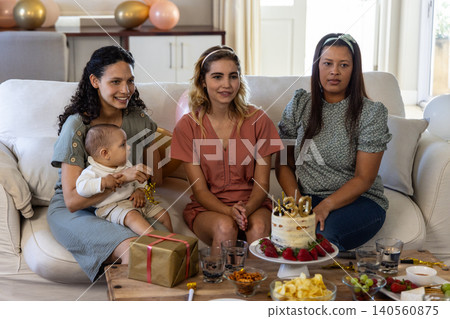 Diverse mothers with baby and friend sitting on couch celebrating birthday with cake and gift box Diverse mothers with baby and friend sitting on couch celebrating birthday with cake and gift box 140560875