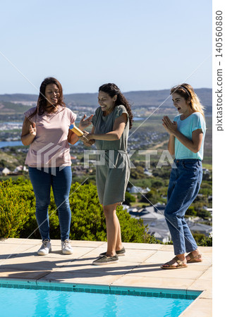 Diverse female friends spraying with water gun on tiled pool deck by pool overlooking coast 140560880
