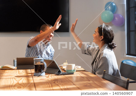 Diverse coworkers wearing business attire high-fiving across conference table at office, laptop 140560907